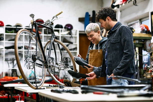 Smiling clerk and customer looking at bicycle seat Smiling female owner with male customer looking at bicycle seat in workshop. Sales clerk is standing with man in bicycle shop. They are in repair shop. bike workshop stock pictures, royalty-free photos & images