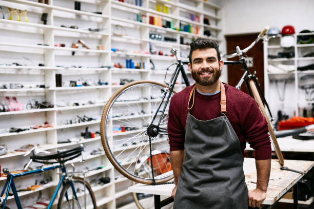 Smiling young bearded owner leaning on workbench Portrait of smiling bearded owner leaning on workbench. Confident young male employee is wearing apron. He is working at bicycle shop. bicycle shop stock pictures, royalty-free photos & images