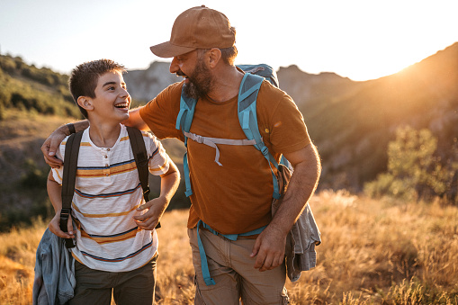 Cheerful teenage boy walking in nature with his dad, embracing, talking and smiling