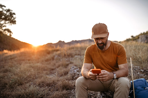 Mature man sitting and taking a break during hiking adventure, using his smart phone