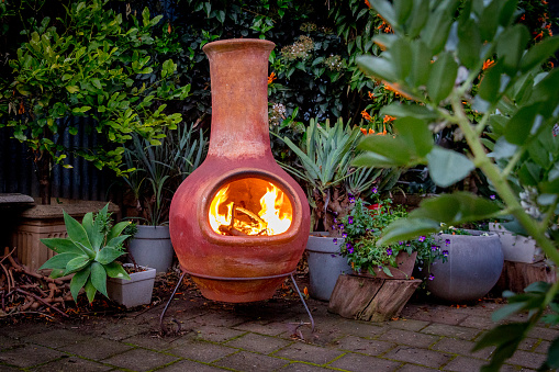 A Chimenea with fire in the backyard with lots of plants in a well landscaped garden.
