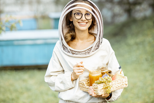 Portrait of a cheerful female beekeeper in protective uniform standing with fresh honey and sweet fruits on the apiary