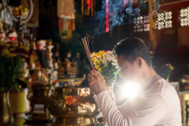Photo of Faith and religious. 
Man hands holding joss sticks praying and blessing in front of altar table at chinese shrine with blurred gods statue in background.