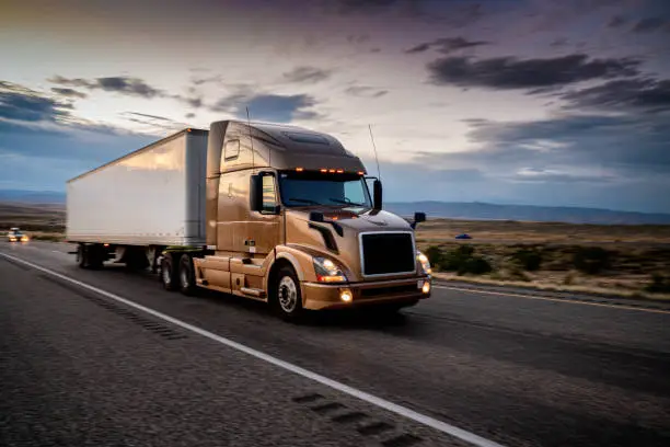 White Semi-Truck Speeding down a four-lane highway with a dramatic sunset in the background and headlights on