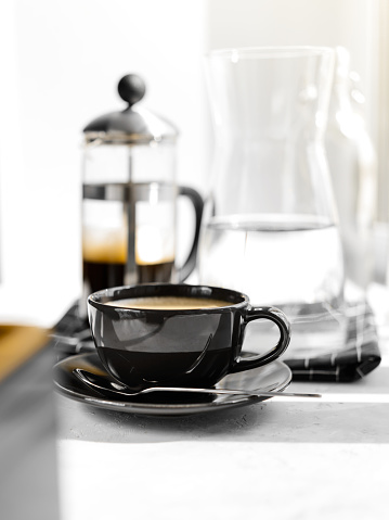 Coffee in black Cup on white background, table. French press, carafe of water, napkin, sunlight. Breakfast.