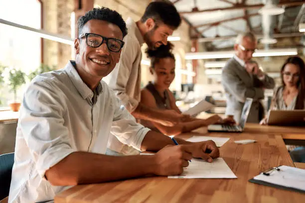 Young and cheerful afro american man in eyeglasses looking at camera with smile while working with his colleagues in the modern office. Business concept. Job