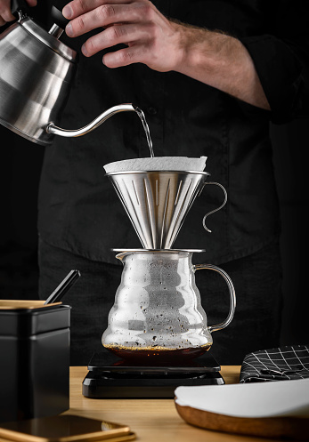 A barista brews coffee by an alternative method in pour over, coffee filter, glass teapot on a wooden tray on a dark background.