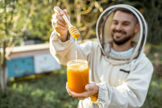 Beekeeper with honey on the apiary Portrait of a handsome beekeper in protective uniform standing with honey in the jar, tasting fresh product on the apiary outdoors apiculture photos stock pictures, royalty-free photos & images