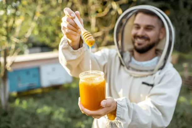Portrait of a handsome beekeper in protective uniform standing with honey in the jar, tasting fresh product on the apiary outdoors