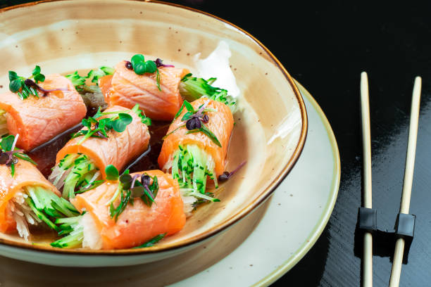 istock Close up on creative made sushi roll with salmon, microgreen, cucumber. Delicious salmon sashimi in yellow bowl with soy sauce on dark background. Japanese cuisine. Seafood, raw fish. 1173187336