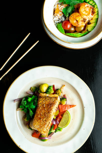 istock Composition with two dishes on dark background. Pike perch fillet with vegetables and Buddha bowl with shrimp and avocado. Delicious wholesome food for diet. Seafood. Food photo for recipe or menu 1173187394