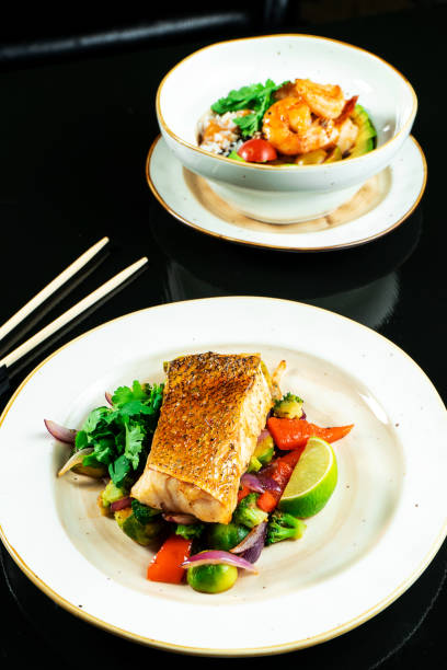 istock Composition with two dishes on dark background. Pike perch fillet with vegetables and Buddha bowl with shrimp and avocado. Delicious wholesome food for diet. Seafood. Food photo for recipe or menu 1173187398