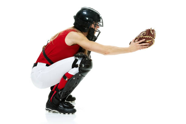 Full length / side view / profile view of 20-29 years old adult beautiful brown hair / long hair caucasian female / young women baseball catcher / athlete / softball player crouching / squatting / bending in front of white background wearing baseball glove / catcher's mask who is serious / confidence / concentration who is catching / throwing with eye black and holding softball - ball / playing softball - sport / baseball - sport / sport longitud completa / vista lateral / vista de perfil de 20-29 años adulto hermoso cabello castaño / pelo largo caucásico hembra / joven mujeres receptor de béisbol / atleta / jugador de softball agachado / en cuclillas / flexión frente al fondo blanco