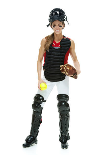 Full length / front view of 20-29 years old adult beautiful brown hair / long hair caucasian female / young women baseball catcher / athlete / softball player standing in front of white background wearing baseball glove / catcher's mask who is serious / confidence / concentration who is catching with eye black and holding softball - ball / playing softball - sport / baseball - sport / sport longitud completa / vista frontal de 20-29 años de edad adulto hermoso cabello castaño / pelo largo caucásico mujer / joven mujeres receptor de béisbol / atleta / jugador de softball de pie frente al fondo blanco usando guante de béisbol / máscara de