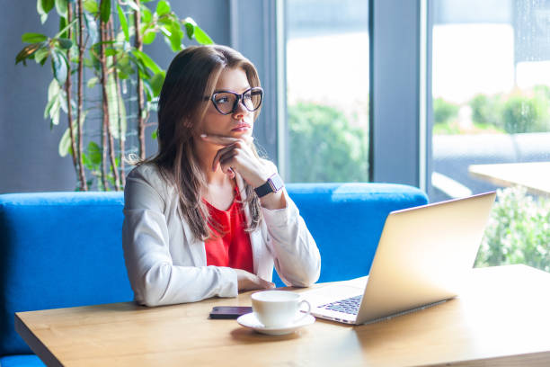 portrait of thoughtful beautiful stylish brunette young woman in glasses sitting with laptop, touching her chin, looking away and thinking what to do - pensive question mark teenager adversity imagens e fotografias de stock