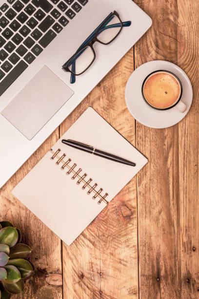 Vertical top view composition of modern office workspace with laptop, note book, coffee and plant on a rustic wooden desk. Office work, study, writing concepts.
