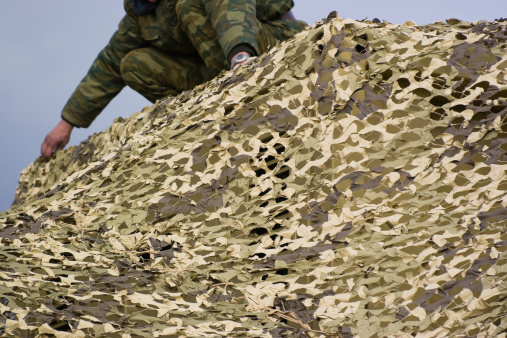 A person in green camouflage sets up a camouflage net with a cloudy sky in the background.Army man prepares a net for military training uses only.