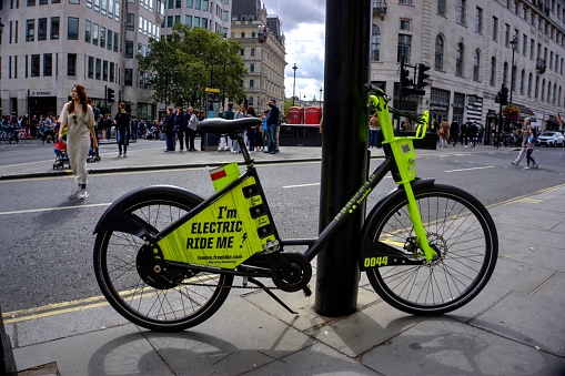 London, United Kingdom - September 7, 2019: Yellow electric assist hire bicycle parked against metal post in central London with tourists in background