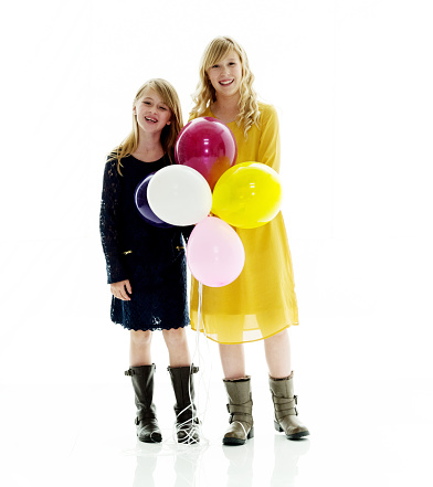 Two people / full length / front view of 8-9 years / 10-11 years old beautiful blond hair / long hair caucasian / generation z young women / female teenage girls / friendship standing in front of white background wearing dress / boot who is smiling / happy / cheerful / cool attitude and celebration and holding balloon / helium balloon / celebration event