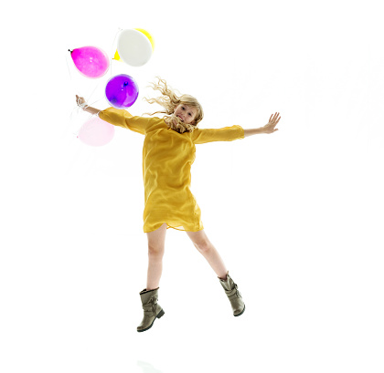 Full length / front view / looking at camera of 10-11 years old beautiful blond hair / long hair caucasian / generation z young women / female teenage girls jumping in front of white background wearing dress / boot who is smiling / happy / cheerful / cool attitude and celebration and holding balloon / helium balloon / celebration event