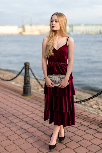 Beautiful slender blonde girl in elegant burgundy velvet ankle-length dress looking away on the summer embankment. Vertical.