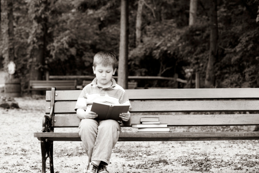 Black and white image. School age boy reading in a park setting.