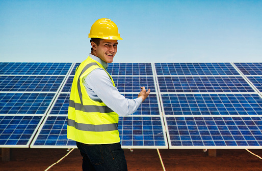 One man only / waist up / side view / profile view / looking at camera of 20-29 years old adult handsome people caucasian male / young men building contractor / construction worker / electrician / engineer / technician installing / standing at the solar power station who is outdoors wearing reflective clothing / waistcoat / protective workwear / jacket / helmet / work helmet / hardhat / button down shirt / shirt who is smiling / happy / cheerful who is working and supporting alternative energy / environmental conservation / renewable energy / solar energy / solar panel / sustainable resources