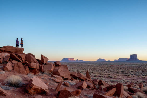 夜明けにアリゾナ州モニュメントバレーの有名なナバホ部族公園で広大な砂漠と赤い岩の風景を楽しむ伝統的な衣服の2つのティーンエイジネイティブネイティブインディアンナバホ姉妹 - people child twin smiling ストックフォトと画像