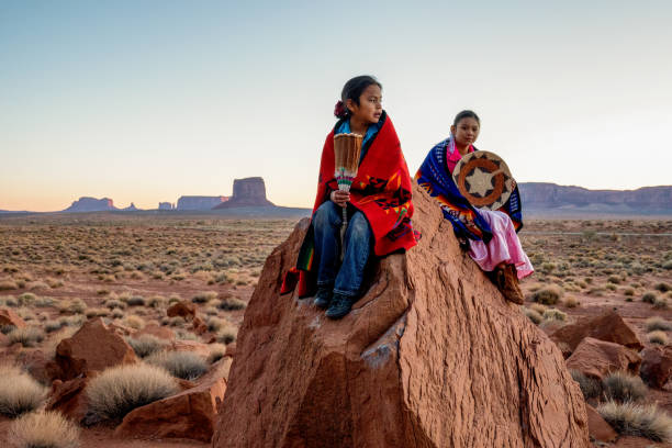 giovane fratello e sorella navajo nella monument valley in posa su rocce rosse di fronte alle incredibili formazioni rocciose di mittens nel deserto all'alba - cherokee foto e immagini stock