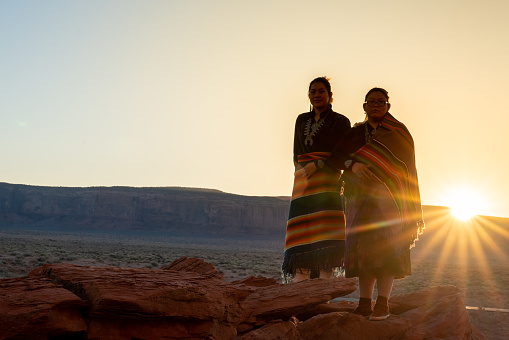 Two Teenage Native American Indian Navajo Sister in Traditional Clothing Enjoying the Vast Desert and Red Rock Landscape in the Famous Navajo Tribal Park in Monument Valley Arizona at Dawn