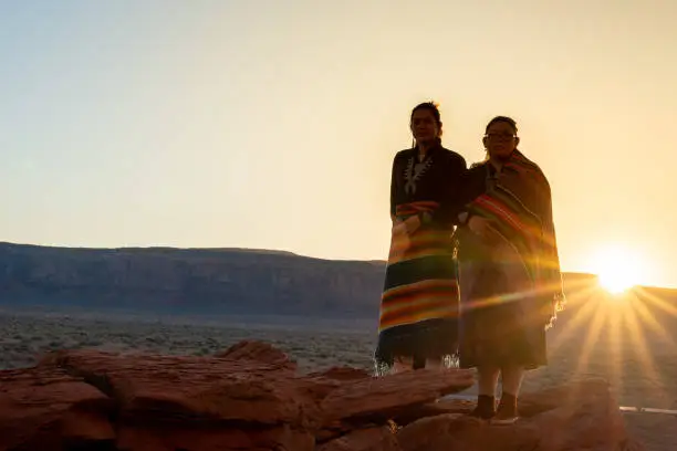 Two Teenage Native American Indian Navajo Sister in Traditional Clothing Enjoying the Vast Desert and Red Rock Landscape in the Famous Navajo Tribal Park in Monument Valley Arizona at Dawn