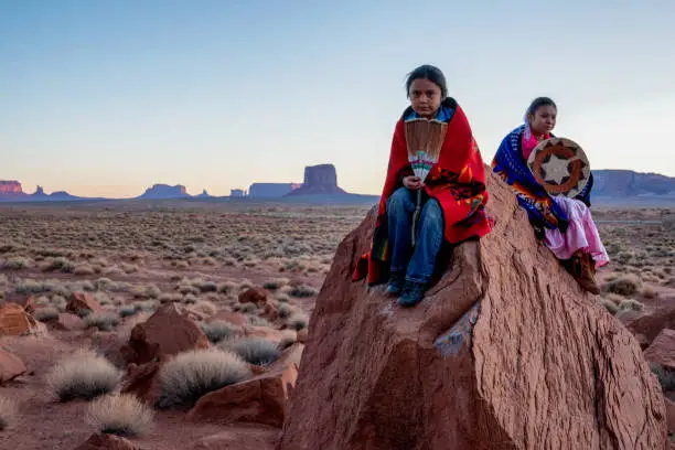 Photo of Young Navajo Brother and Sister in Monument Valley Posing on Red Rocks in front of the Amazing Mittens Rock Formations in the Desert at Dawn