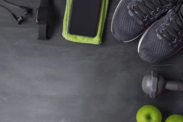 Photo of Fitness healthy lifestyle and weight loss concept. Top view of athlete's equipment  dumabbell, smartphone, smart watch, sport shoes and green apple on black background.