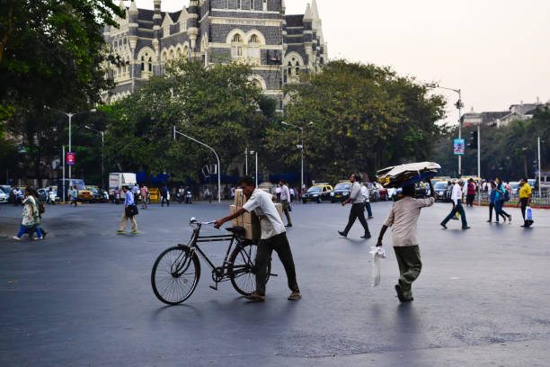 Mumbai, Maharashtra, India - December, 2016: People crossing road on pedestrian. Man carrying boxes on his bicycle. Other man hiding from rain under cardboard boxes