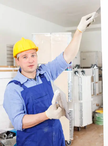 Confident male plasterer working at indoors building site, renovating wall with spatula and plaster