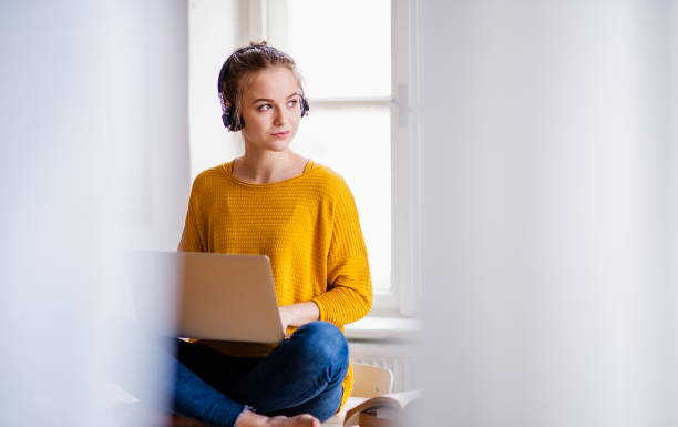 une jeune étudiante s'asseyant sur le bureau, utilisant des écouteurs en étudiant. - working at home audio photos et images de collection