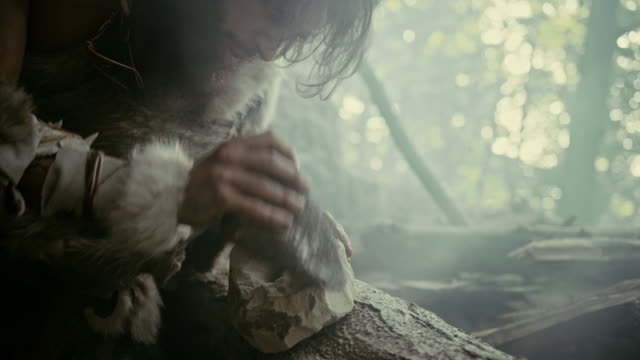 Primeval Caveman Wearing Animal Skin Hits Rock with Sharp Stone, Makes First Primitive Tool for Hunting Animal Prey. Neanderthal Using Flint Rock. Dawn of Human Civilization. Slow Motion Closeup Shot
