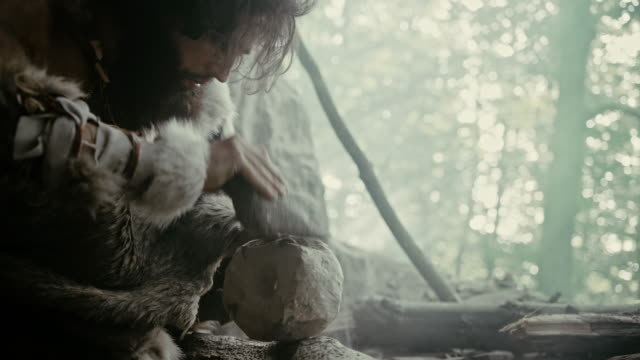 Primeval Caveman Wearing Animal Skin Hits Rock with Sharp Stone and Makes Primitive Tool for Hunting Animal Prey. Neanderthal Using Hand axe to Create first Wheel. Slow Motion Shot