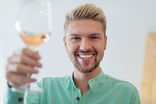 Blond young man with stubble beard and pierced chin rising glass of white wine and smiling