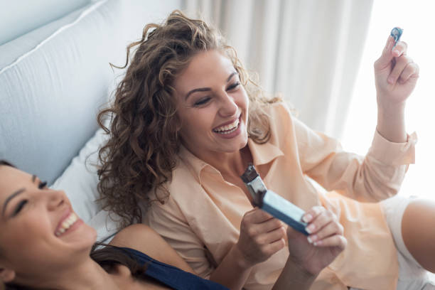 Two young women eating chocolate bars in bed and laughing