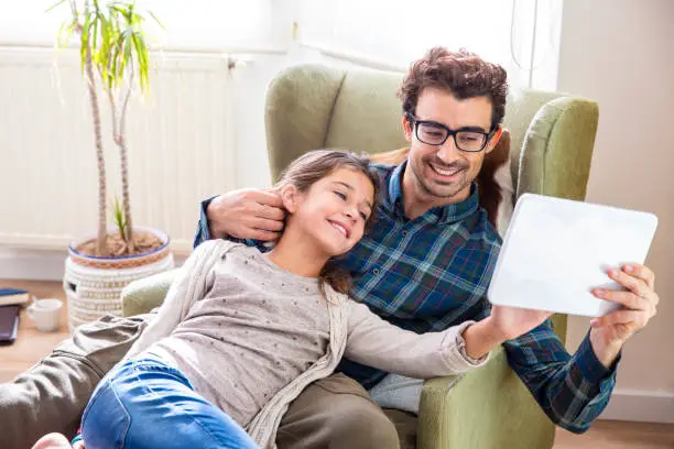 father and daughter in armchair playing tablet pc in living room