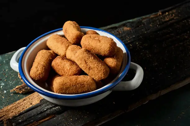 closeup of some spanish croquettes in a white and blue enamel plate on a rustic dark gray wooden table, against a black background