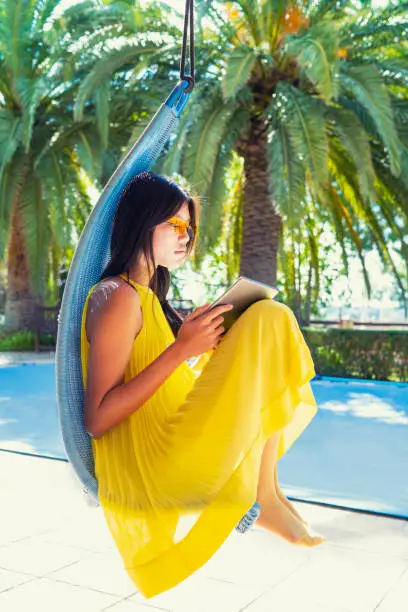 Asian woman reading tablet on swing chair with yellow dress in backyard with palm trees