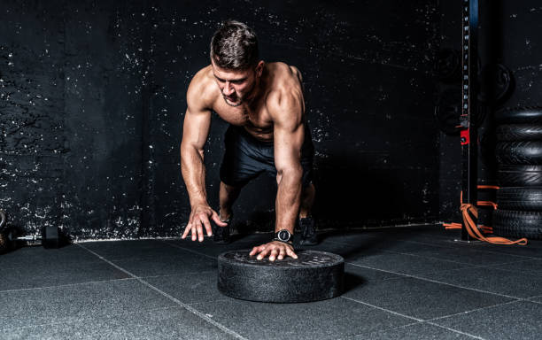 istock Young strong sweaty focused fit muscular man with big muscles doing push ups with one hand on the barbell weight plate and jump for training hard core workout in the gym real people selective focus 1173340121