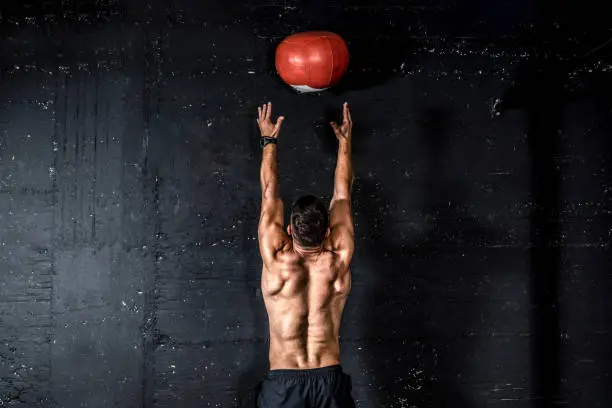 Med ball training, Young strong sweaty focused fit muscular man with big muscles doing throwing medicine ball up on the wall for training hard core workout in the gym real people selective focus