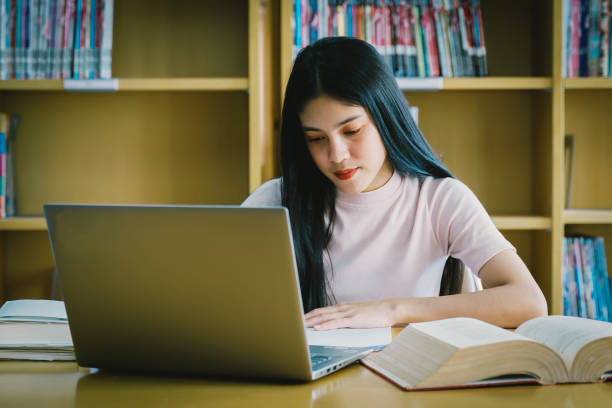 istock Young Asian woman student study and take notes with book in library. Female university student sitting at table doing assignments in college library. Student using laptop and learning online. 1173361732