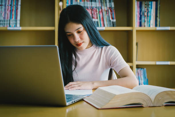 istock Young Asian woman student study and take notes with book in library. Female university student sitting at table doing assignments in college library. Student using laptop and learning online. 1173361761