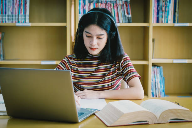istock Young Asian woman student study and take notes with book in library. Female university student sitting at table doing assignments in college library. Student using laptop and learning online. 1173361840