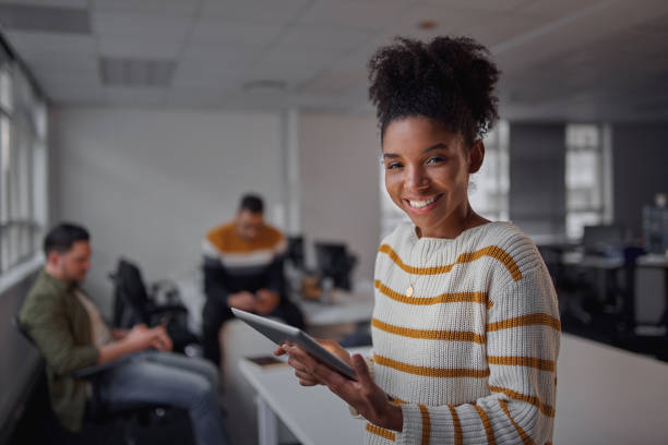 fröhliche junge afrikanische amerikanische geschäftsfrau in casual wear halten digitale tablet lächelnd und blick in die kamera, während ihre kollegen im hintergrund kommunizieren - unternehmerin - frauen empowerment - mädchen chef - defocused business office casual stock-fotos und bilder