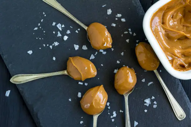 Photo of Caramel in spoons and in bowl close up on black background with salt flakes - top view